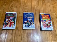 Three Disney VHS tapes with Christmas themes laid out on wooden floor: Beauty and the Beast: The Enchanted Christmas, Mickey's Magical Christmas, and Winnie the Pooh and Christmas Too.