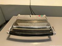 Top view of closed sandwich press showing stainless steel lid, black handle, and power indicator buttons with attached power cord.