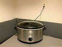 Stainless steel Crock-Pot slow cooker with glass lid. Lid knob missing revealing metal screw. Black control knob on front. Plugged in and placed on a corner surface.