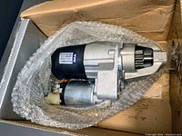 Starter motor lying in bubble wrap inside an open box. Metal body with black and silver colors, visible label showing brand TYF.