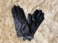 Pair of new black leather gloves lying on a cream shaggy surface, showing front and back sides with tags visible.