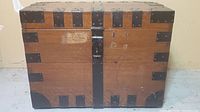 Front view of the antique wooden trunk showing metal corner reinforcements, central latch, and worn paper label.