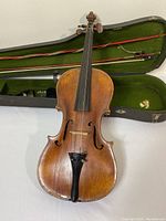Front view of the violin laid on a white surface. Violin shows wooden craftsmanship with aged patina and slight wear marks. Violin case visible in background with green interior lining. Two violin bows placed inside the case.