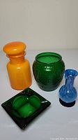 Four colored glass pieces: orange jar with lid, green jar with embossed design, blue vase, and green square dish