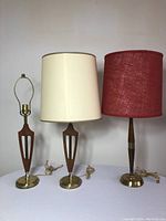 Three Mid-Century Modern style table lamps together: two with off-white drum shades and one with red fabric shade, bases wood and brass.