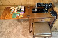 Full view of Singer 15J sewing machine mounted in wooden sewing table with fold-out leaf extended, cushioned bench in front, and various sewing accessories on table surface.