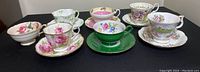 Seven vintage teacup and saucer sets arranged in two rows on a black surface. Variety of colors and patterns including floral and solid green