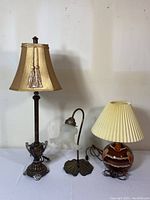 Full view of three vintage lamps on table: tall brass with fabric shade and tassels, small brass with frosted glass shade on leaf base, round pottery with cream pleated shade.