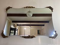 Front view showing full rectangular beveled glass mirror with ornate brass-coloured Victorian style decorative metal accents on four corners and center top and bottom lengths, hung on wall reflecting room.