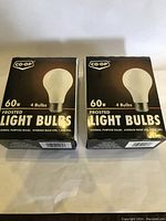 Two sealed boxes each containing 4 frosted 60 watt Co-Op light bulbs.