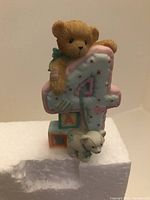 Close-up of a teddy bear figurine holding pink and white number 4 with ABC blocks and small white dog at base.
