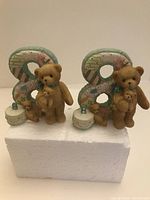 Two Cherished Teddies collectibles with teddy bears holding smaller bears, each paired with a pastel number 8 and a small cake with candle, displayed on white foam block