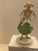 Figurine of a woman in a green dress holding a child in white, standing on a white base signed 'enjoy me' by Claire Stoner.