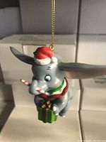 Photo shows a close-up of the vintage Disney Dumbo Christmas tree ornament hanging, depicting Dumbo wearing a red Santa hat, holding a green gift with a candy cane and scarf.