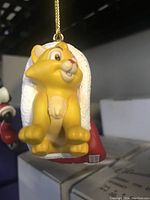 Front side of the ornament showing the yellow kitten character Oliver sitting on a white textured base with red accents and a gold hanging cord.