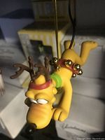 Photo showing Disney Pluto Christmas tree ornament in yellow with brown reindeer antlers and red collar with gold bells, hanging with original box visible behind.