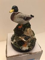 Front and side view of mallard duck figurine on original white box showing the ducks perched on a sculpted natural base resembling a log with greenery.