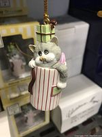 Close-up of the gray cat ornament with red-striped container and green present, showing detail and gold hanging string.