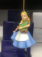 Photo of a vintage Alice in Wonderland Christmas tree ornament featuring Alice in a blue dress and white apron holding a decorated green tree with stars, hanging from a gold string.