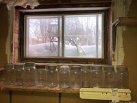 Twelve clear glass canning jars with metal lids displayed in a basement window area with snow view outside.