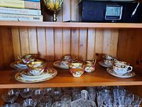 Wide view of shelf showing rose-pattern teacups, saucers and creamers on wooden backdrop