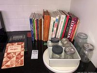 Photo showing a range of cookbooks from vintage to modern, glass canning jars, and plastic containers on a kitchen counter.