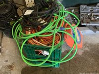 Bin filled with green, orange, black, and white extension cords coiled and stored together in a green crate next to tools and a power strip.