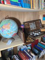 Photo of a colorful world globe on a beige stand next to an open wooden box with chess pieces inside, with books on shelves below.