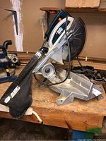 Side view of Black & Decker miter saw showing overall unit with dust collection bag attached, placed on a wooden workbench.