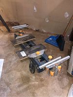 Full view of portable miter saw stand showing metal frame with adjustable support arms, orange adjustment knobs, and wheels for mobility on concrete floor.