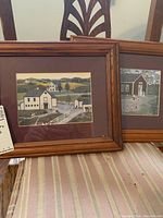 Two wooden framed prints with rural farm scenes leaning against chairs. Scenes include houses, barns, fences, and animals with maroon mats.