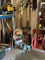 Overall view of yard tools and equipment leaning against a wall including string trimmers, pole saw, push brooms, and other tools.
