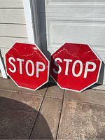 Pair of red reflective STOP signs leaning against a white wall and garage door on a concrete surface outdoors.