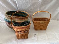 Three woven baskets shown together on a white cloth background: Longaberger basket, a tall multicolor basket, and a large round basket.