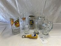 Collection of seven assorted beer glasses and a yellow beer bottle opener, visible on a white backdrop.