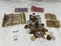 Collection of foreign currency notes and assorted coins laid out on a white cloth surface with a MaxSold tag labeled 65.
