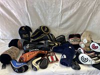 Photo showing an assortment of golf club head covers from multiple brands in various colors and materials, including some golf-related accessories in original packaging.