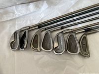 Photo showing heads of seven golf irons from various brands such as Callaway, Cleveland and Wilson Staff with visible club numbers and marks indicating use.