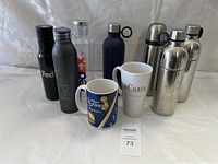 Photo showing assortment of 7 insulated bottles and 2 ceramic mugs on white cloth background.