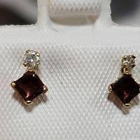 Close-up photo of the pair of earrings showing the deep red square-cut tourmaline and small round diamond above each stone in yellow gold setting.