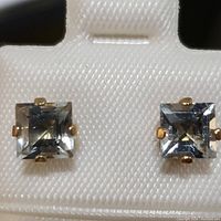 Close-up photo of the pair of square-cut aquamarine earrings in yellow gold prong setting on a white plastic display card.