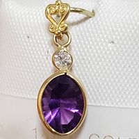 Close-up photo of oval amethyst and diamond pendant in 14K yellow gold, showing purple amethyst and small diamond above with decorative bail.