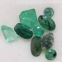 Nine small natural emerald stones in various shapes and cuts, showing natural inclusions and green color variations.