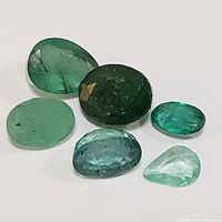 Six loose emerald gemstones arranged on white background showing varying green hues and cuts