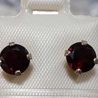Close-up photo of a pair of round faceted deep red garnet stud earrings set in four-prong sterling silver settings on white textured display backing.