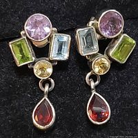 Close-up of left silver earring showing multiple faceted gemstones: round amethyst, rectangular aquamarine, rectangular peridot, round citrine, and pear-shaped garnet dangle.