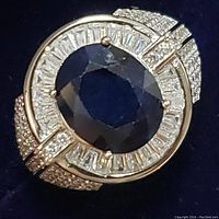Close-up of ring top showing oval dark blue sapphire and surrounding clear baguette stones with encrusted clear round stones on band
