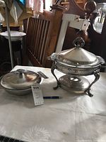 Chafing dish with ornate stand, burner visible, and additional covered casserole dish alongside on table.