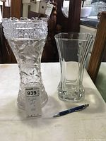 Photo showing two crystal vases on a table with a pen for scale; the taller vase has deep floral and herringbone cuts, the shorter one is square with vertical lines.