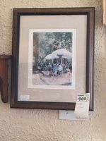 Framed art print showing a cafe scene with people under umbrellas, in a brown wood frame with beige matting.